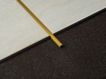 Złota matowa listwa dekoracyjna do płytek ułożona pomiędzy dwiema płytkami na tle brązowego dywanu